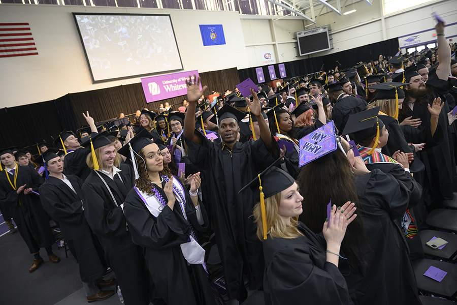 A group of graduates clap at commencement dressed in caps and gowns.