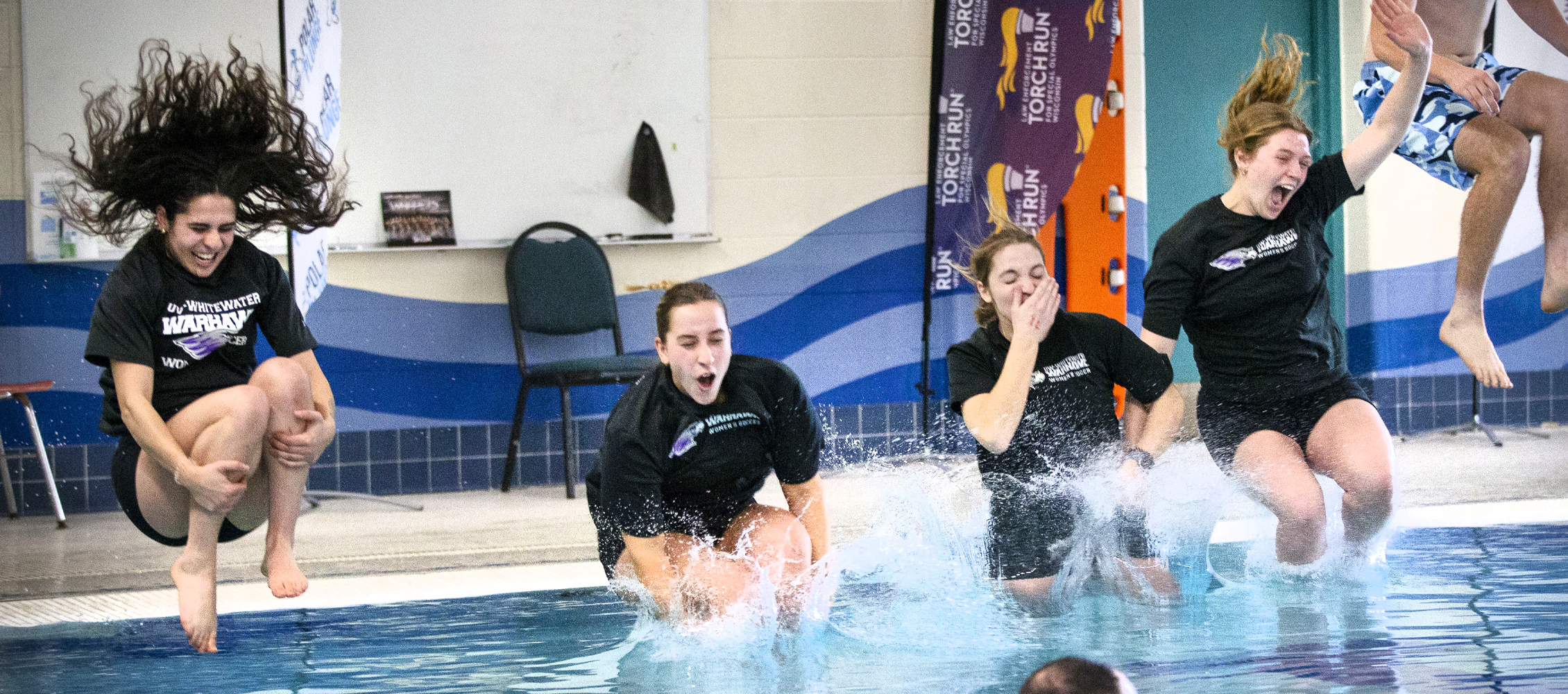 Four people in black shirts jump energetically into an indoor pool.