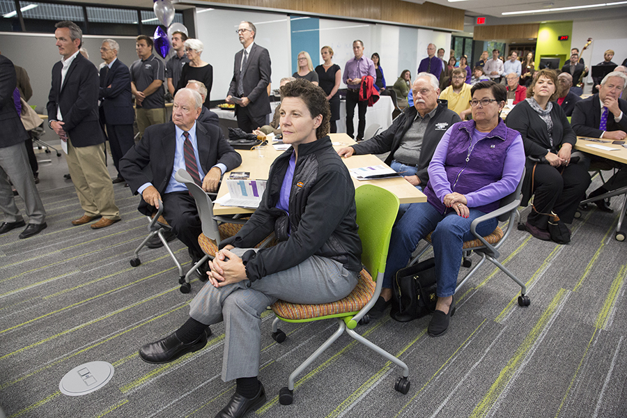 Julie Anding, wearing a black shirt, gray pants and purple tie, sits in a chair at the front of a room full of people with their focus on the front of the room.
