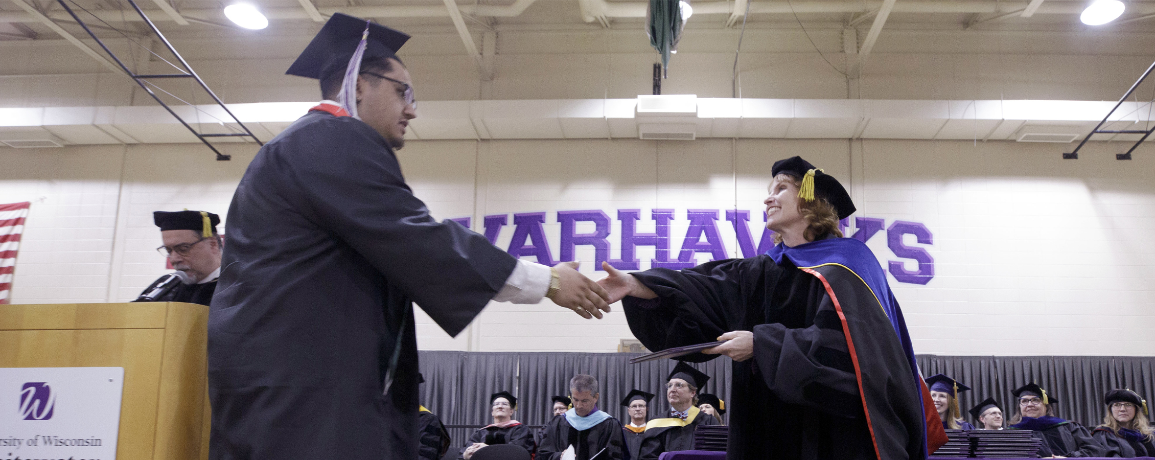 A student wearing academic regalia crosses a stage and shakes hands with Tricia Clasen. 