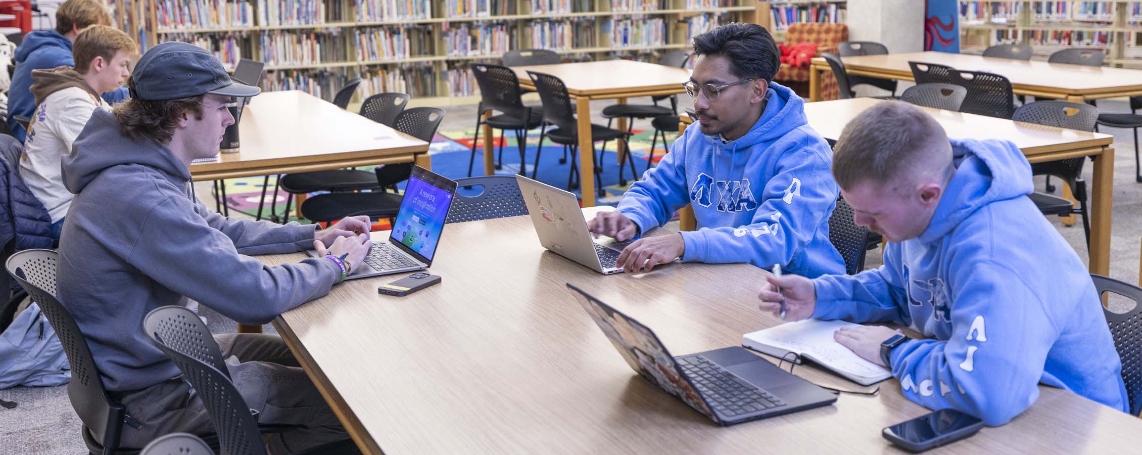 Three male students sitting at table with laptops in library.