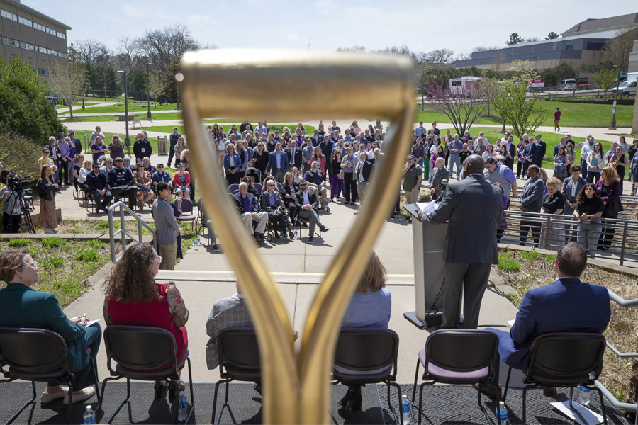Audience members listening to Chancellor Corey A. King at the podium are seen through the handle of a golden ceremonial shovel.