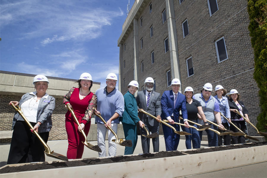 10 people take shovels of dirt in front of building for a ceremonial groundbreaking.