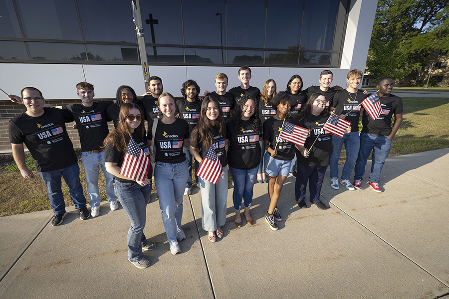 A group of students stand together outdoors holding American flags and wearing black shirts that say Enactus.