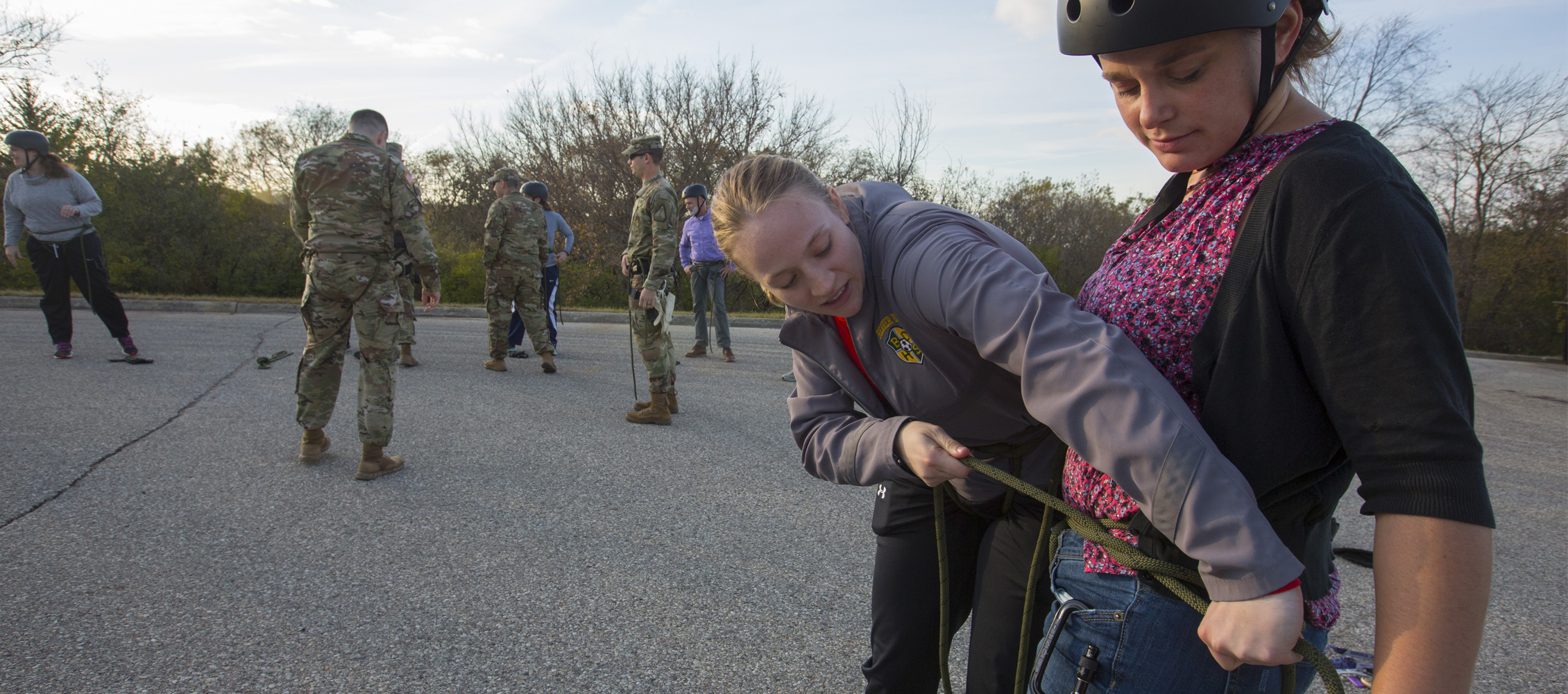 A student helps their instructor tie her harness outside.