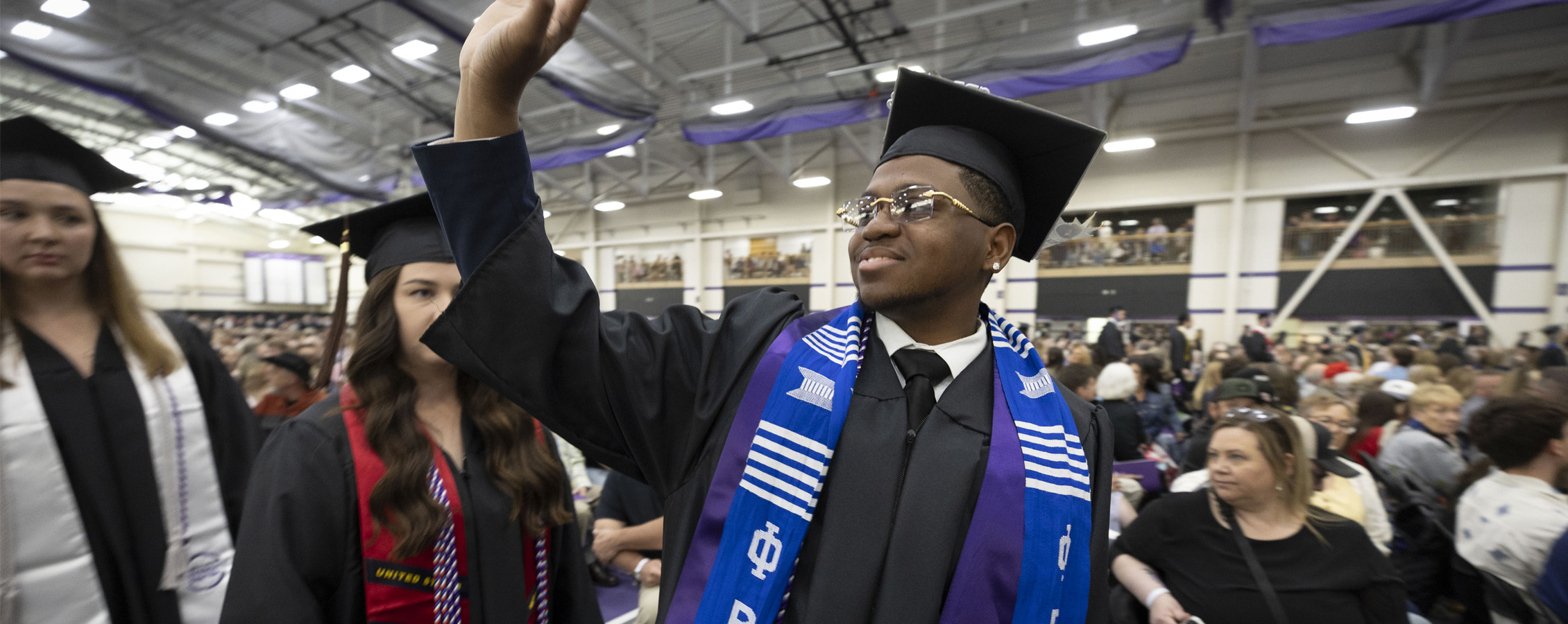 A student in a graduation cap and gown waves to friends and family at commencement ceremony.