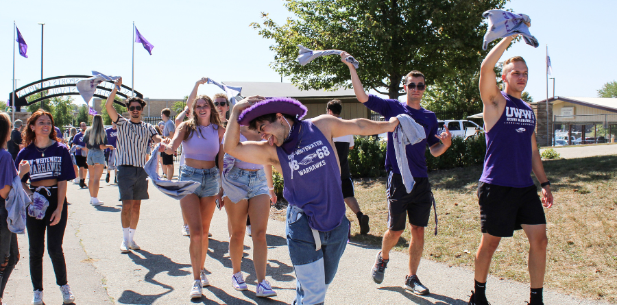 Students walking down a sidewalk wearing purple and waving flags.