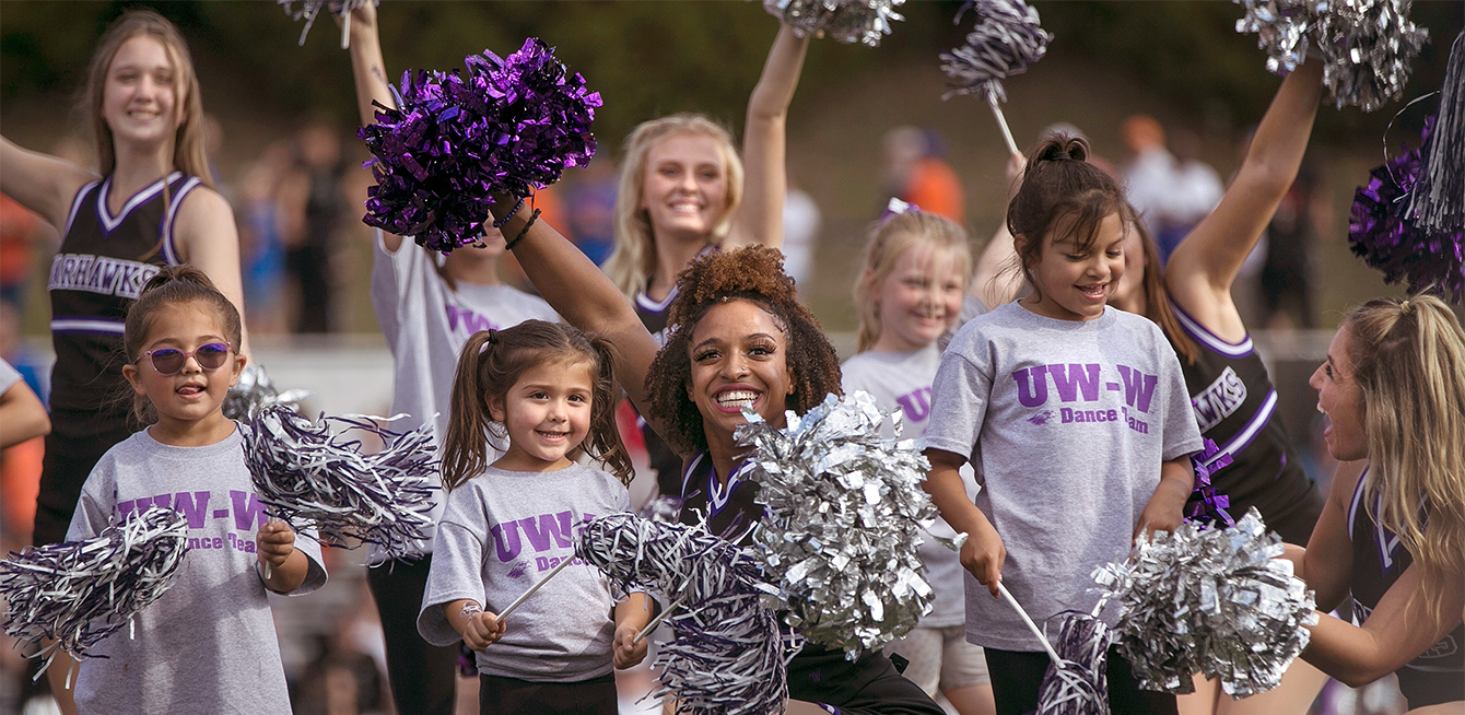 Girls from the poms dance team smile with kids on the football field.