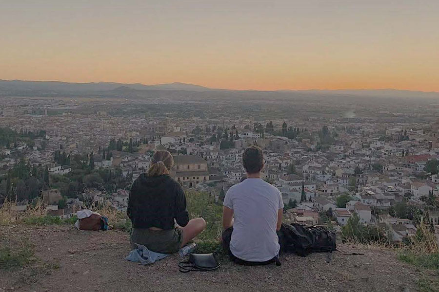 Two people sit on a cliff and look at a city below.