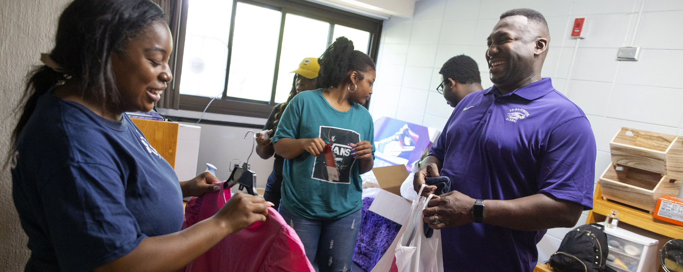 A student holds a pink shirt on a hanger as her family helps her move into her dorm room.