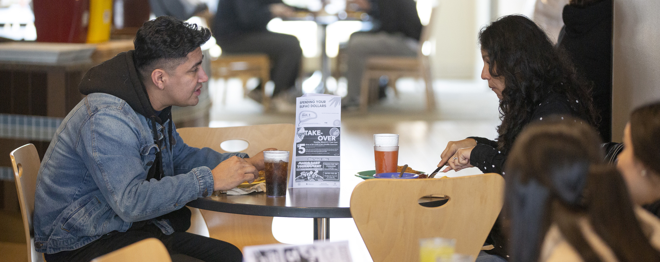 Two people sit at a table together in the dining hall with plates of food in front of them.