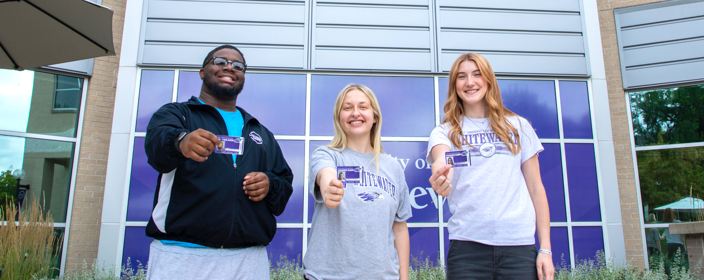Three students stand in front of the University Center, smiling and holding out their HawkCards for the camera.