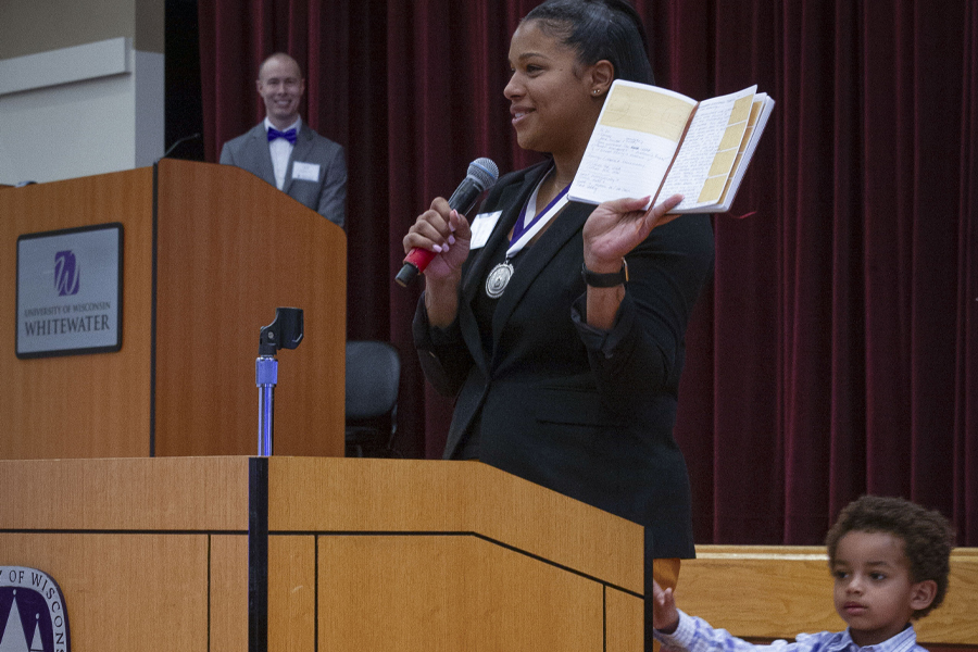 An alum speaks at a podium while holding up a book and a small child stands next to them.