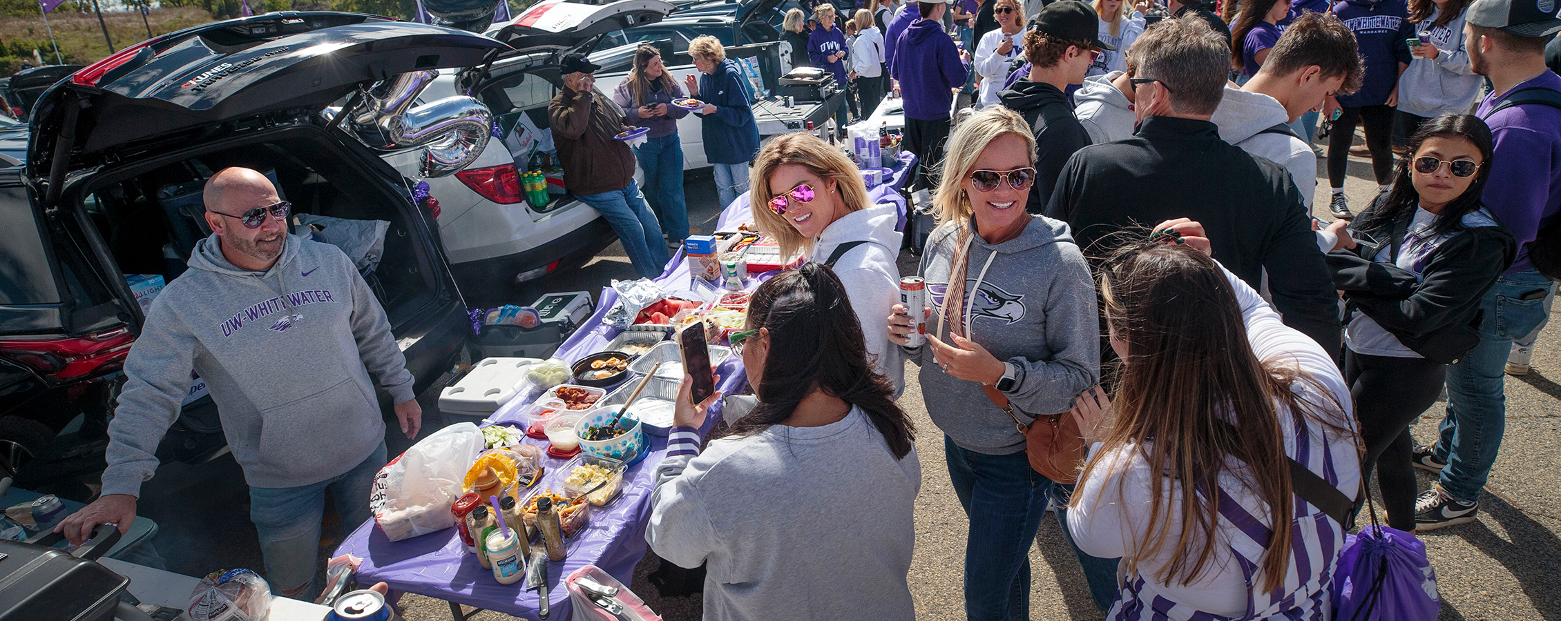 Warhawk fans stand around a long table with food on it while tailgating at a UW-Whitewater football game.