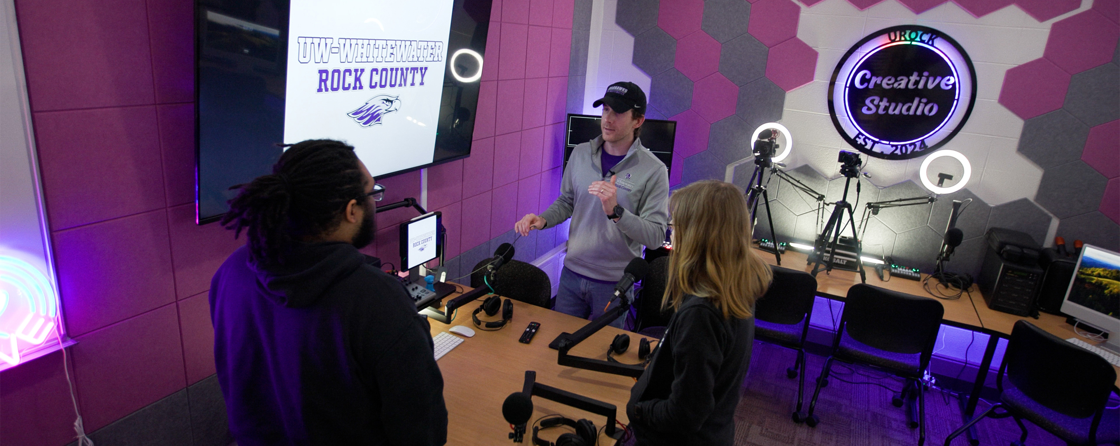 Two students and a faculty member stand in a room with purple walls and a sign that says Creative Studio.