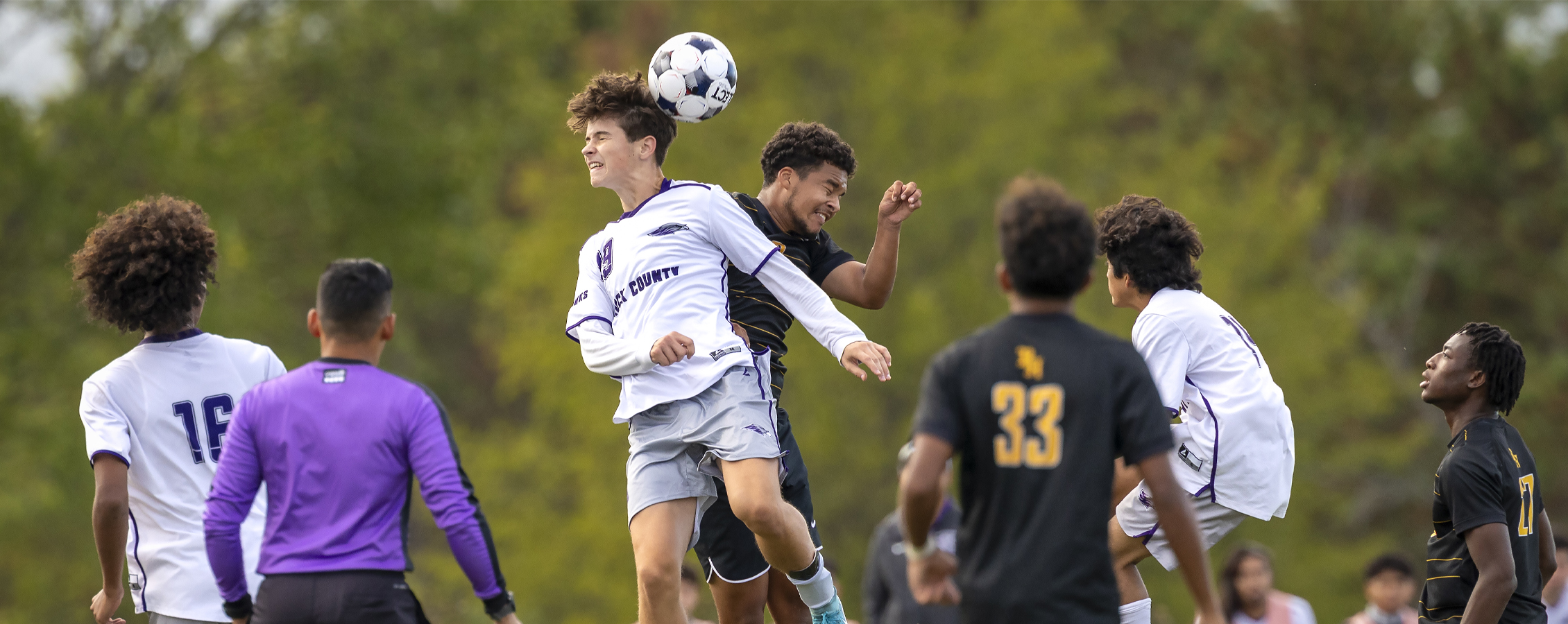 A men's soccer player jumps in the air to head a ball with a competitor jumping right behind him while a ref watches.
