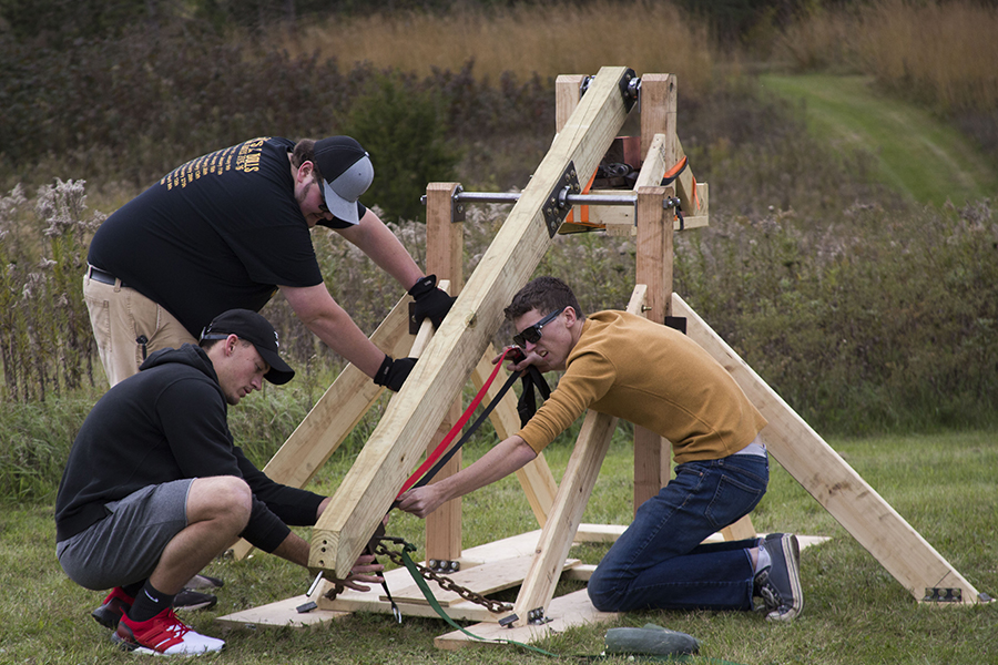 Three students are on the ground outside building a wooden trebuchet.