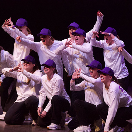 Members of a sorority wear black pants, white shirts, and purple caps and cheer on stage.