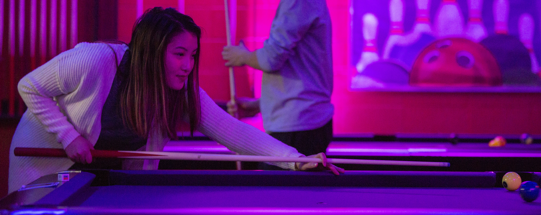 A student leans over a pool table with a pool stick in their hands.