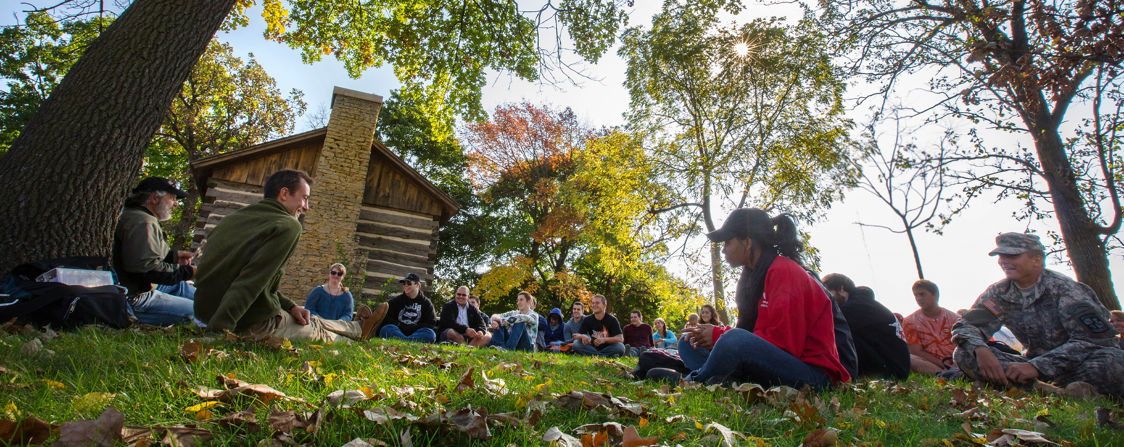  Students sit for a class outdoors with leaves on the ground.