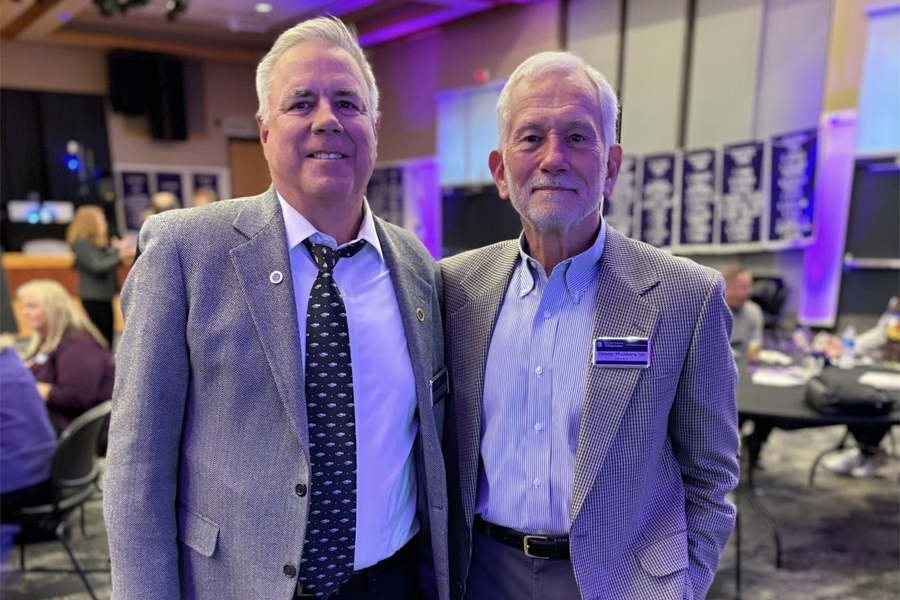 Denny Waldera, wearing a button shirt and blazer, stands in the University Center and poses for a photo with another person.